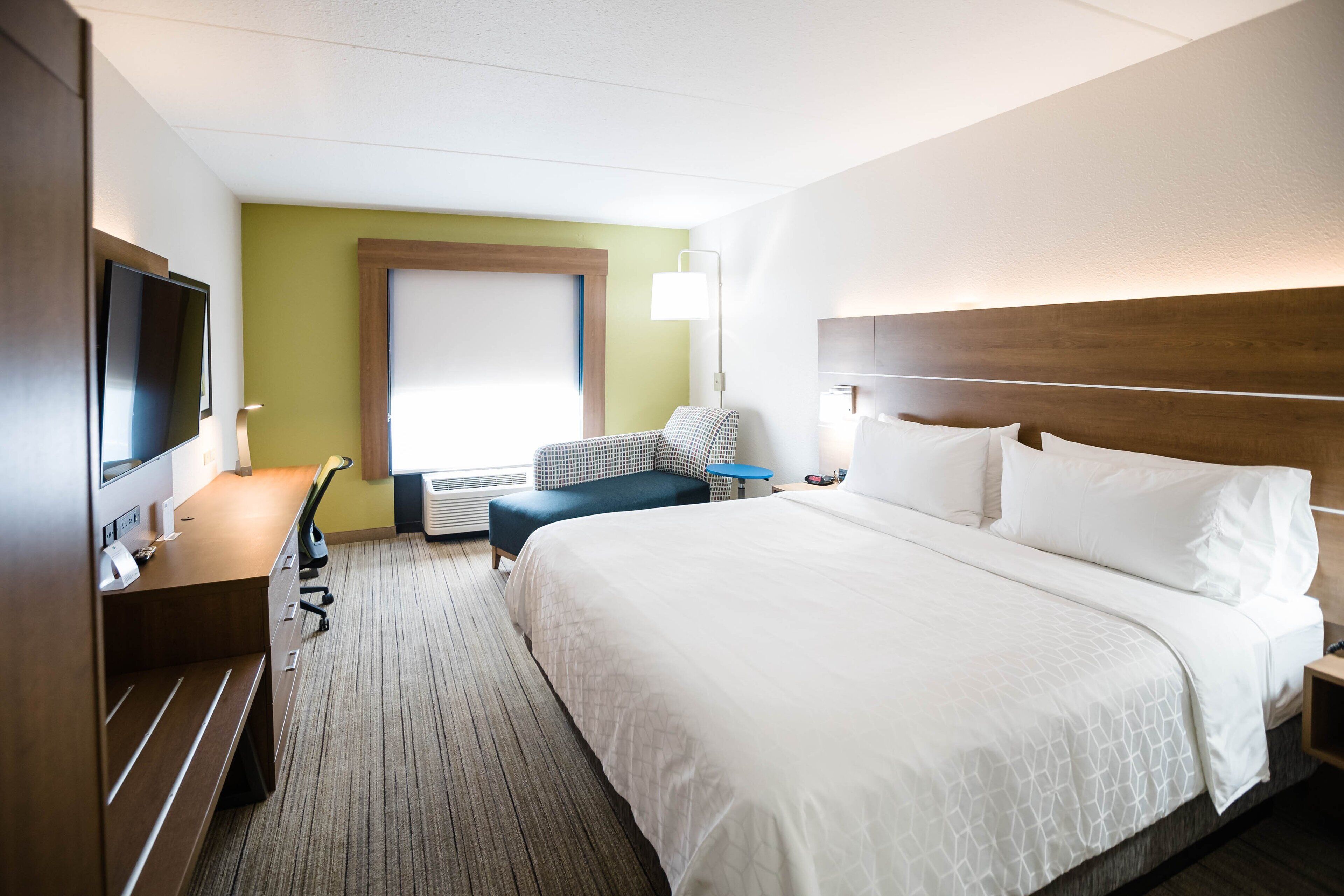 A hotel room with a large bed, a desk with a chair, a television, and a window with blinds.