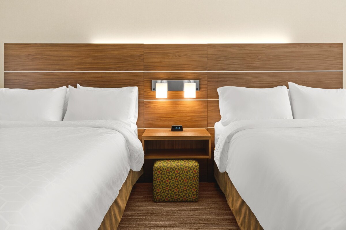A hotel room with two beds, a nightstand, a digital clock, and a patterned ottoman.