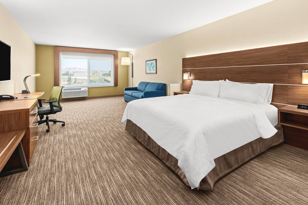 A hotel room with a large bed, a desk with a chair, a TV, a window with a view, and a wall-mounted lamp.