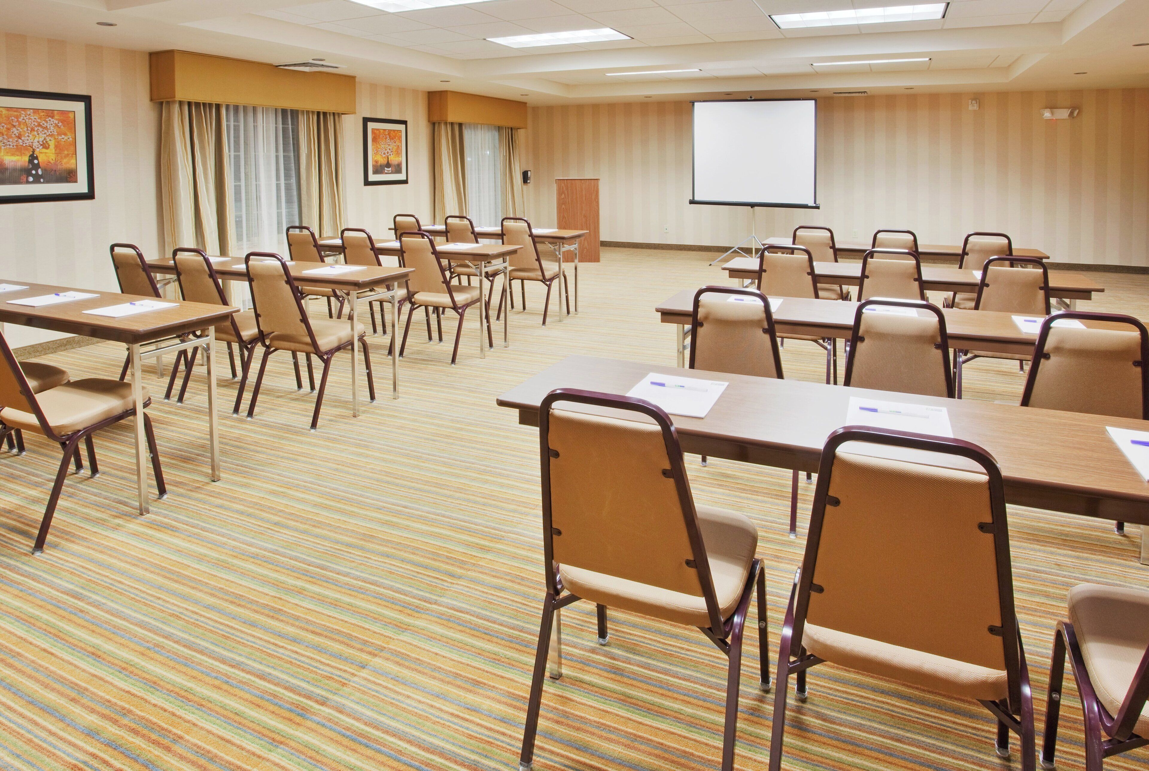 Un salón de conferencias con varias mesas y sillas, una pantalla de proyector y piso alfombrado.
