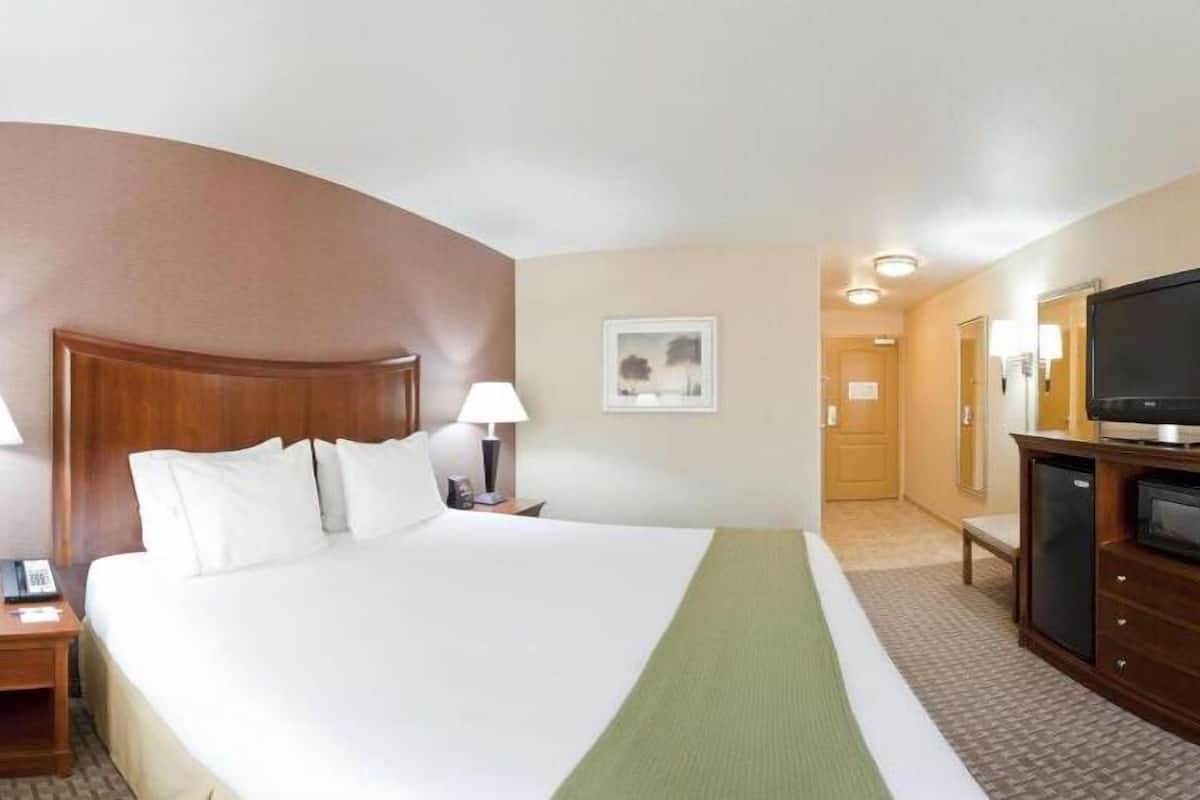 A hotel room with a large bed, two bedside lamps, a flat-screen TV, a minibar, and a door leading to another room.