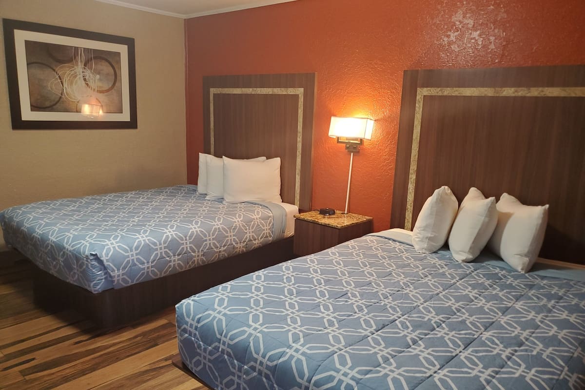 A hotel room with two beds, each with a patterned bedspread and white pillows. There is a nightstand with a lamp between the beds.
