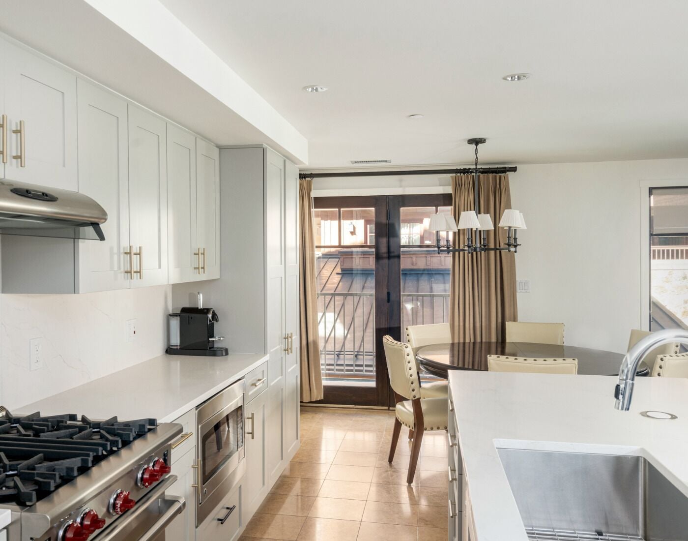 A modern kitchen with white cabinets, a gas stove, an oven, and a coffee maker. There is a dining area with a table and chairs, and a view of a balcony through a glass door.