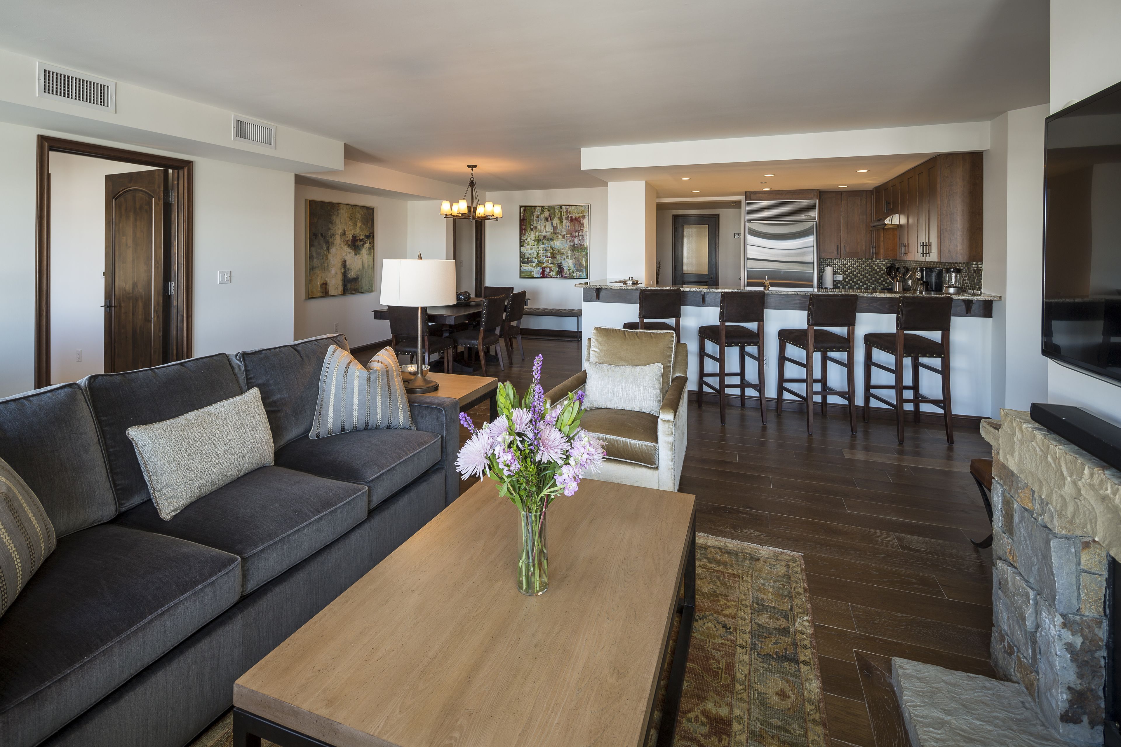 A modern living room with a dark gray sofa, a wooden coffee table with a vase of flowers, and a kitchen in the background with a bar area.
