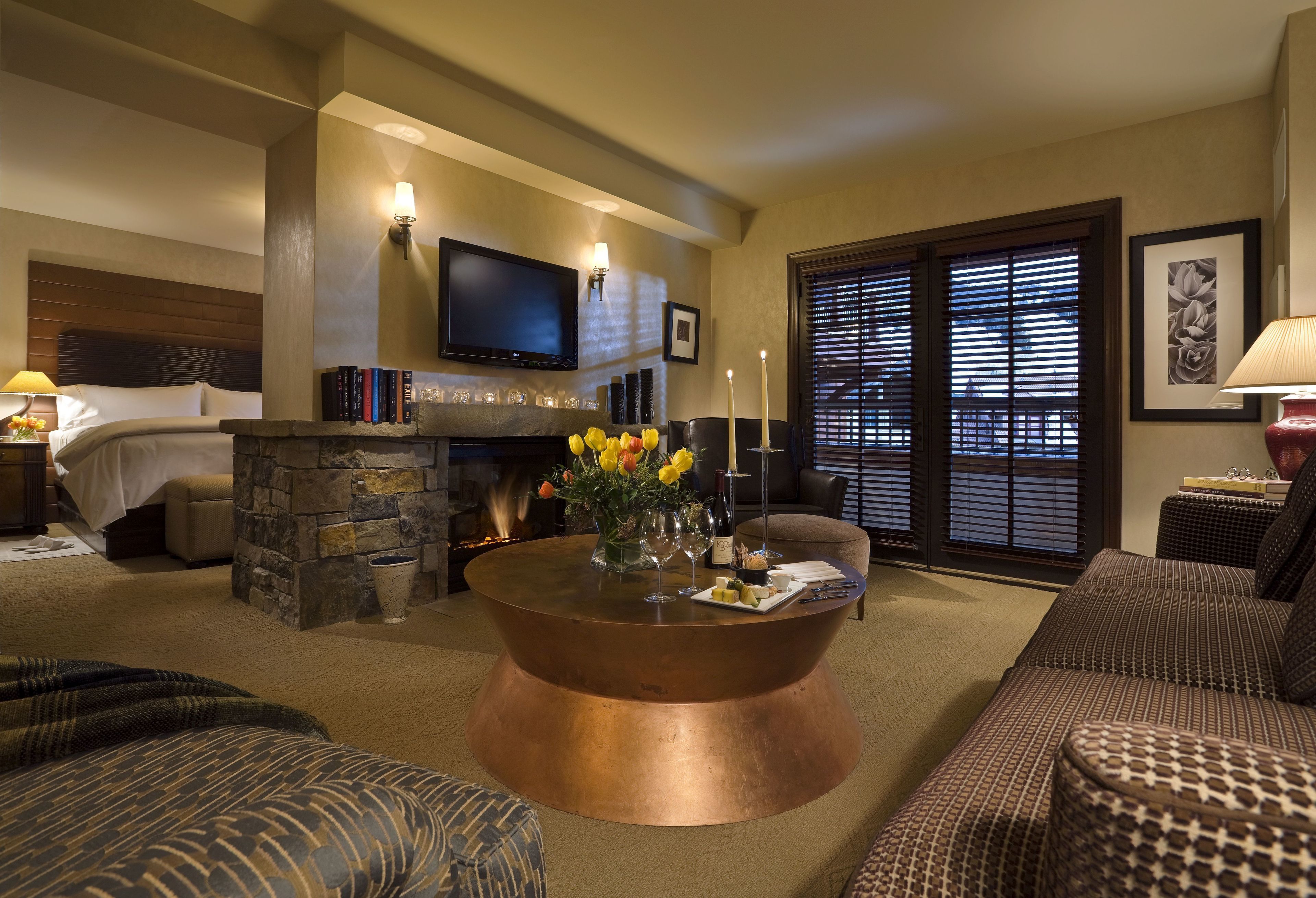 A hotel room with a stone fireplace, a flat-screen TV mounted on the wall, a comfortable sofa, and a coffee table with a vase of flowers.