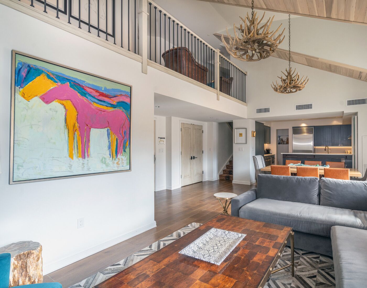 A spacious living area with a large abstract painting, a wooden coffee table, a gray sofa, and a kitchen in the background.