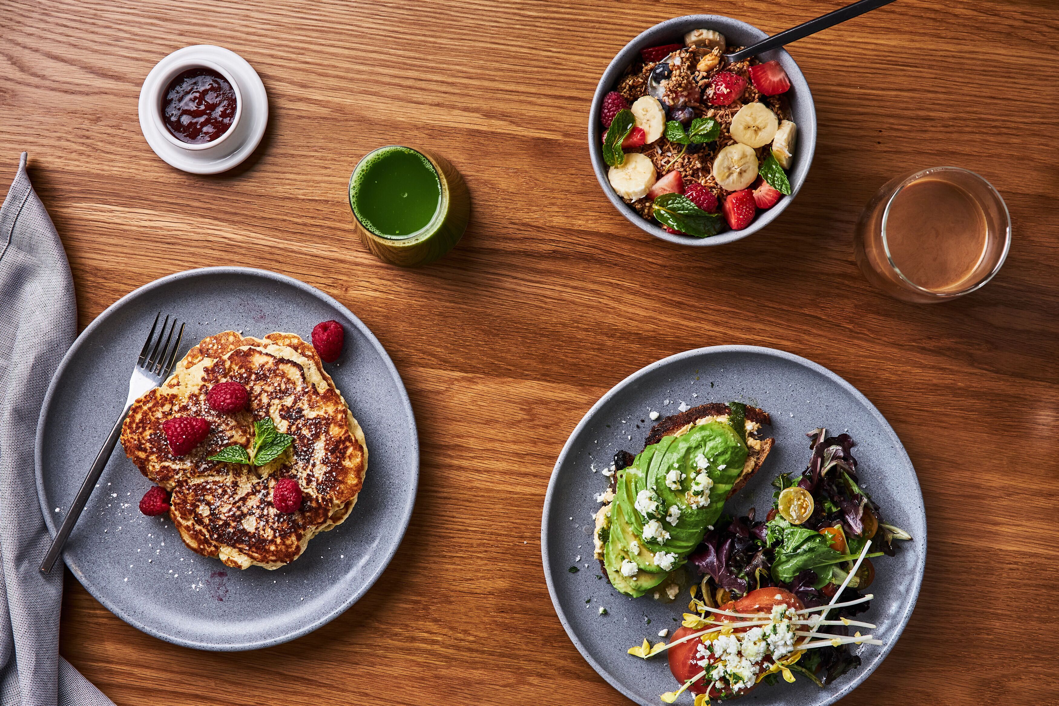 Une table en bois sur laquelle sont disposés des aliments pour le petit-déjeuner, notamment des crêpes, un saladier de fruits et une assiette de toasts à l’avocat.