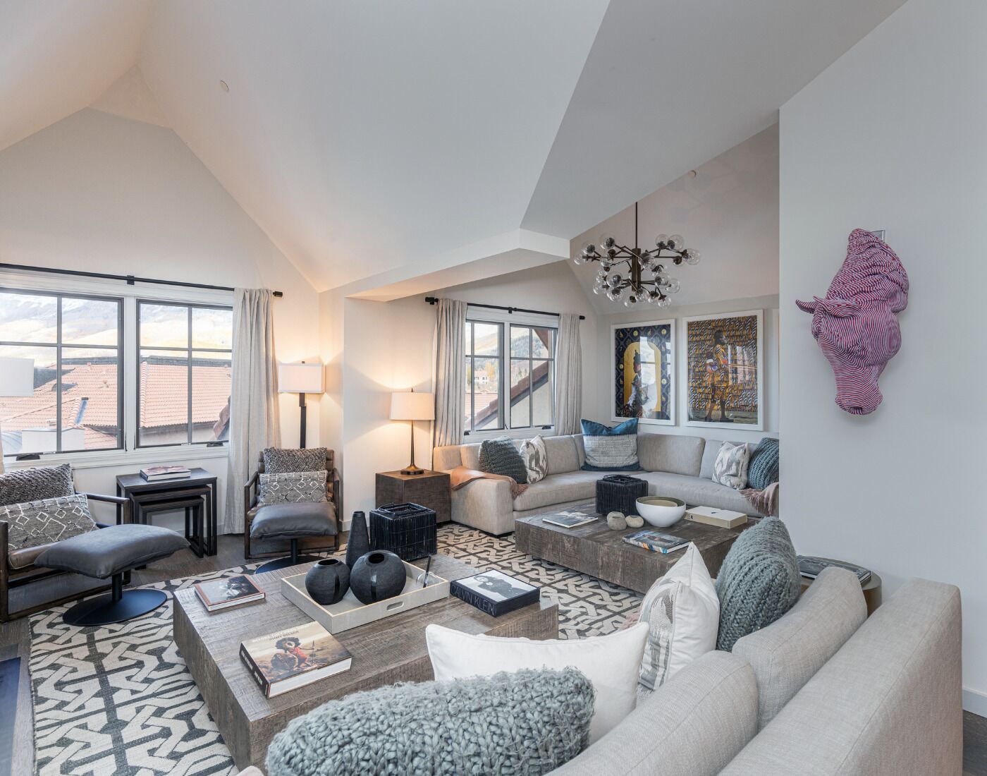 A spacious living room with a large sofa, a coffee table, and a fireplace. There are large windows providing natural light and a view of the outside.