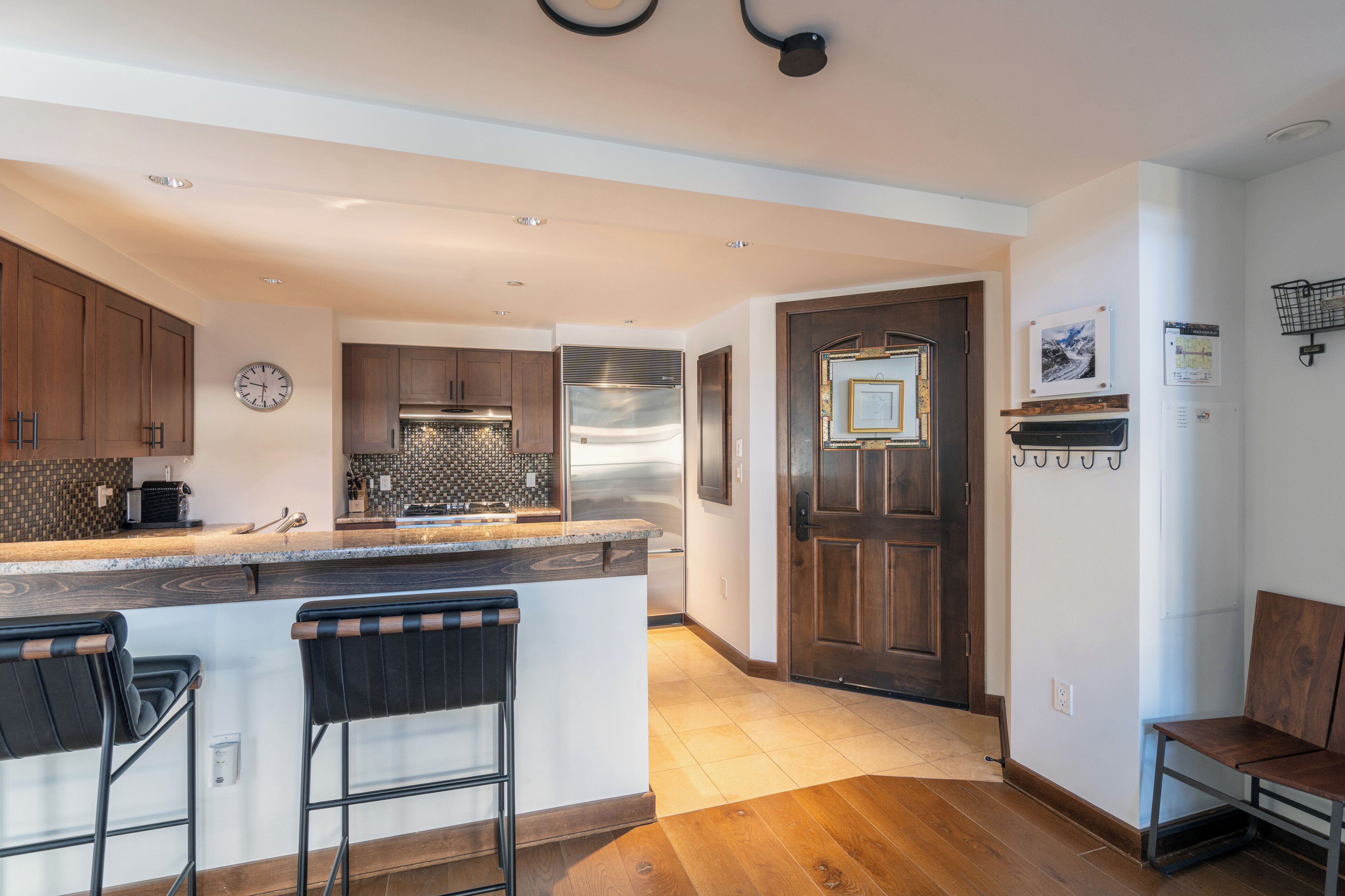 A modern kitchen with a central island, bar stools, wooden cabinets, and a stainless steel refrigerator.