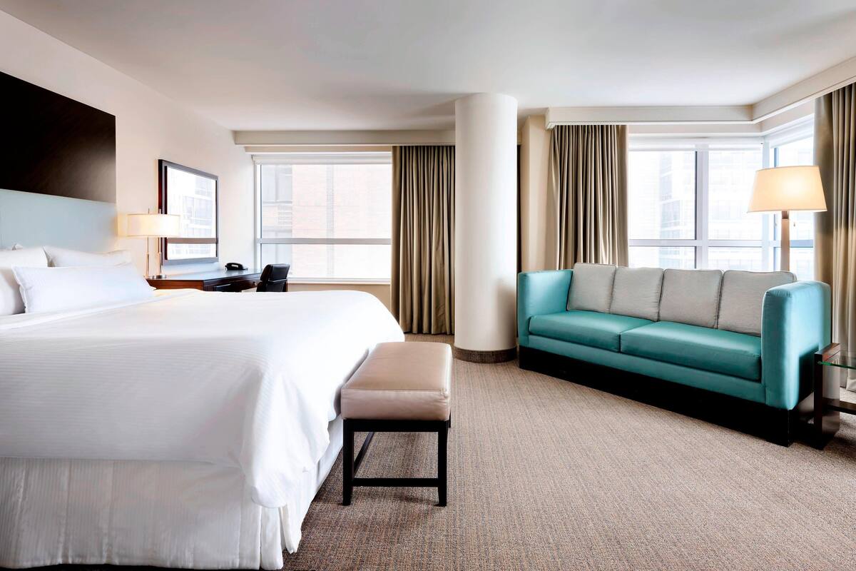 A hotel room with a large bed, a teal sofa, a small table, and a window with curtains.