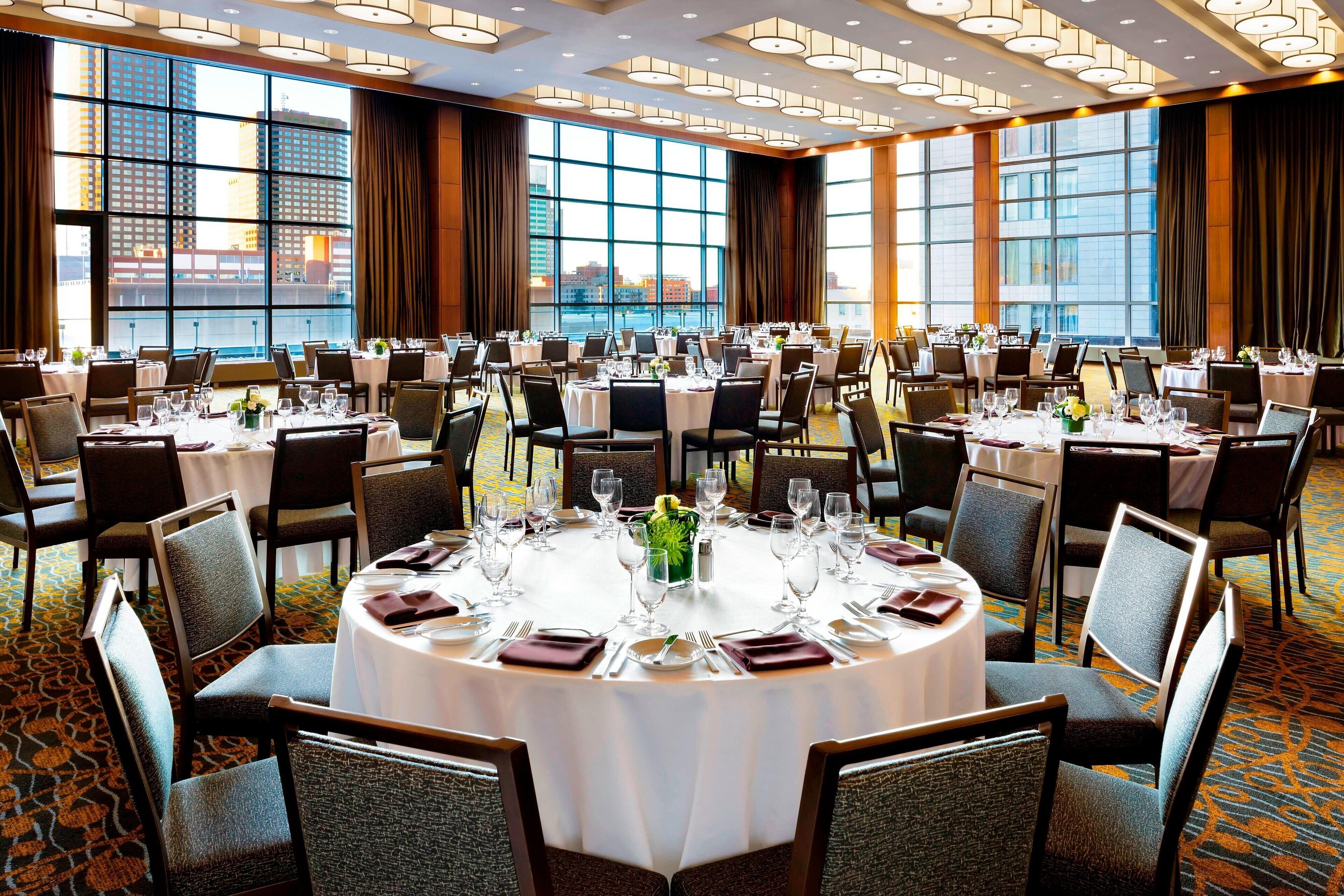 Un amplio salón de banquetes con mesas redondas dispuestas para el servicio de comidas, ventanas grandes con cortinas y vistas a edificios.