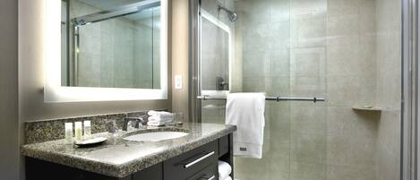 Une salle de bain moderne dotée d’un comptoir en granit, de deux lavabos, d’un grand miroir, d’une douche à parois de verre et d’un porte-serviettes.