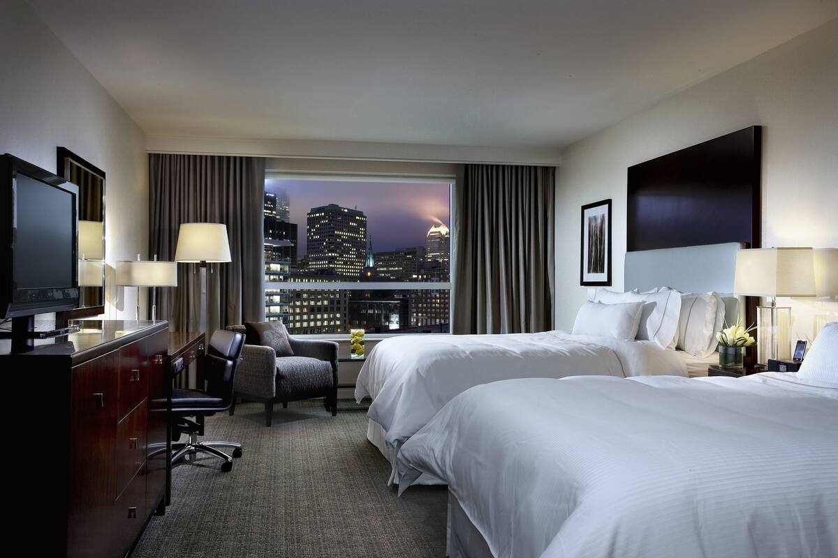 A hotel room with two beds, a desk, a chair, a TV, and a view of a cityscape at night.