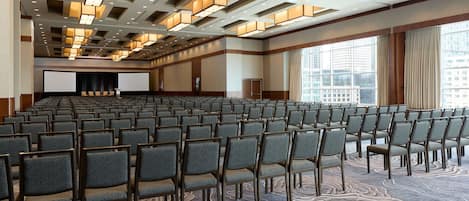 A large conference room with rows of chairs, a stage with a projector, and large windows offering a city view.