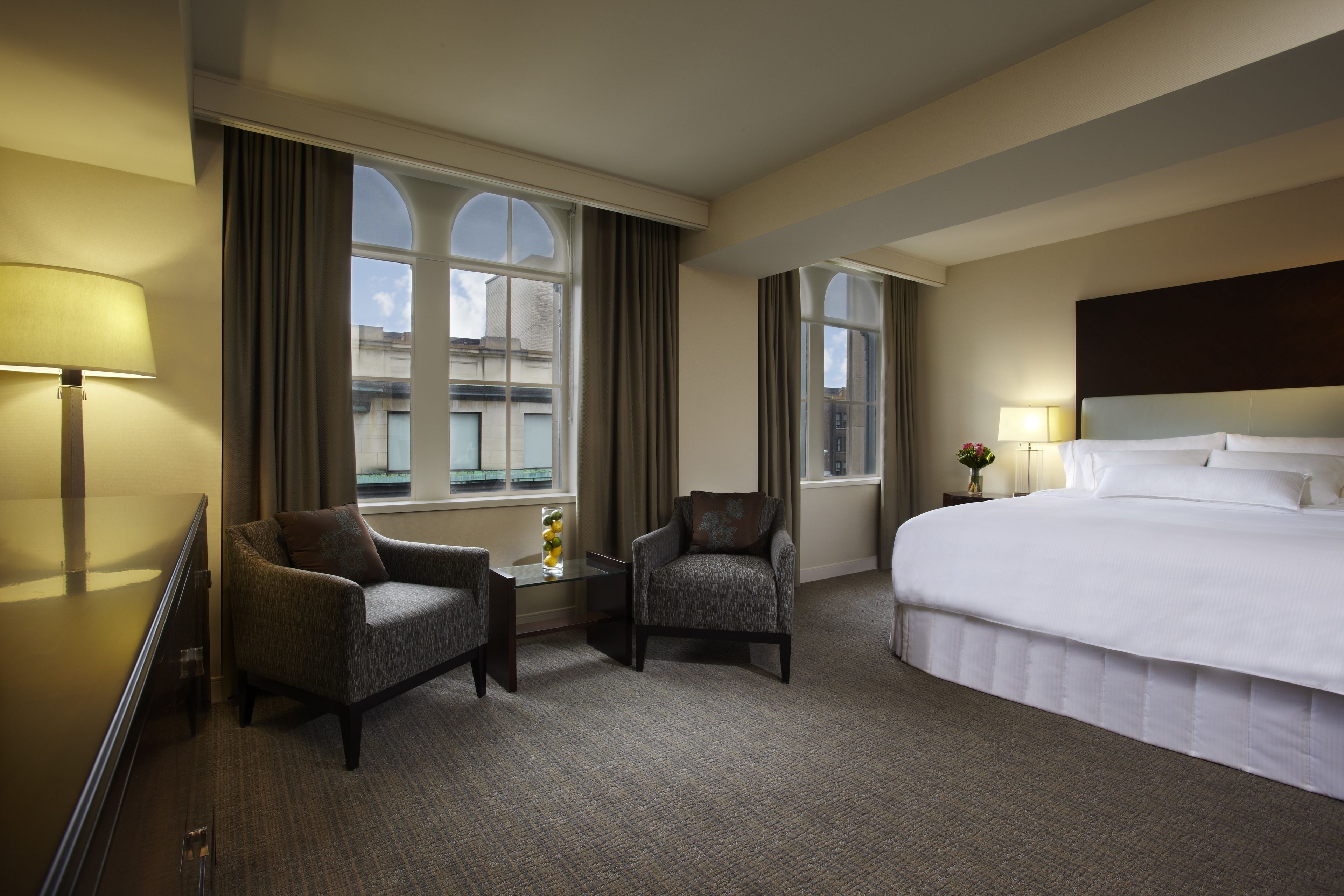 A hotel room with a large bed, two armchairs, a small table with a vase, and large windows with curtains.