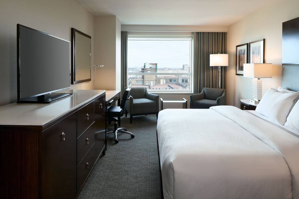 A hotel room with a large bed, a desk with a flat-screen TV, two armchairs, a small table, and a window with a city view.