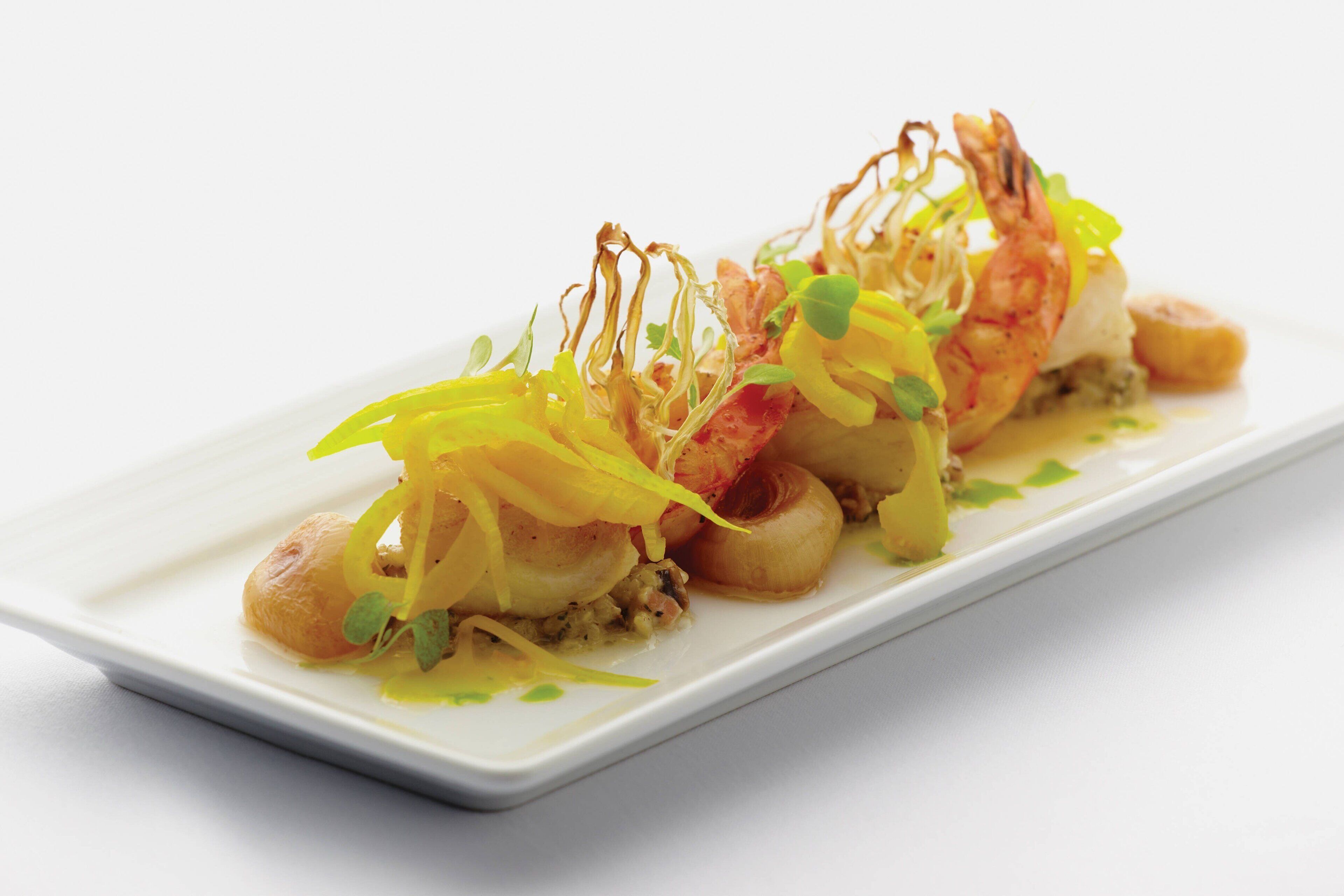 A white rectangular plate with a seafood dish, including shrimp and scallops, garnished with yellow and green elements.