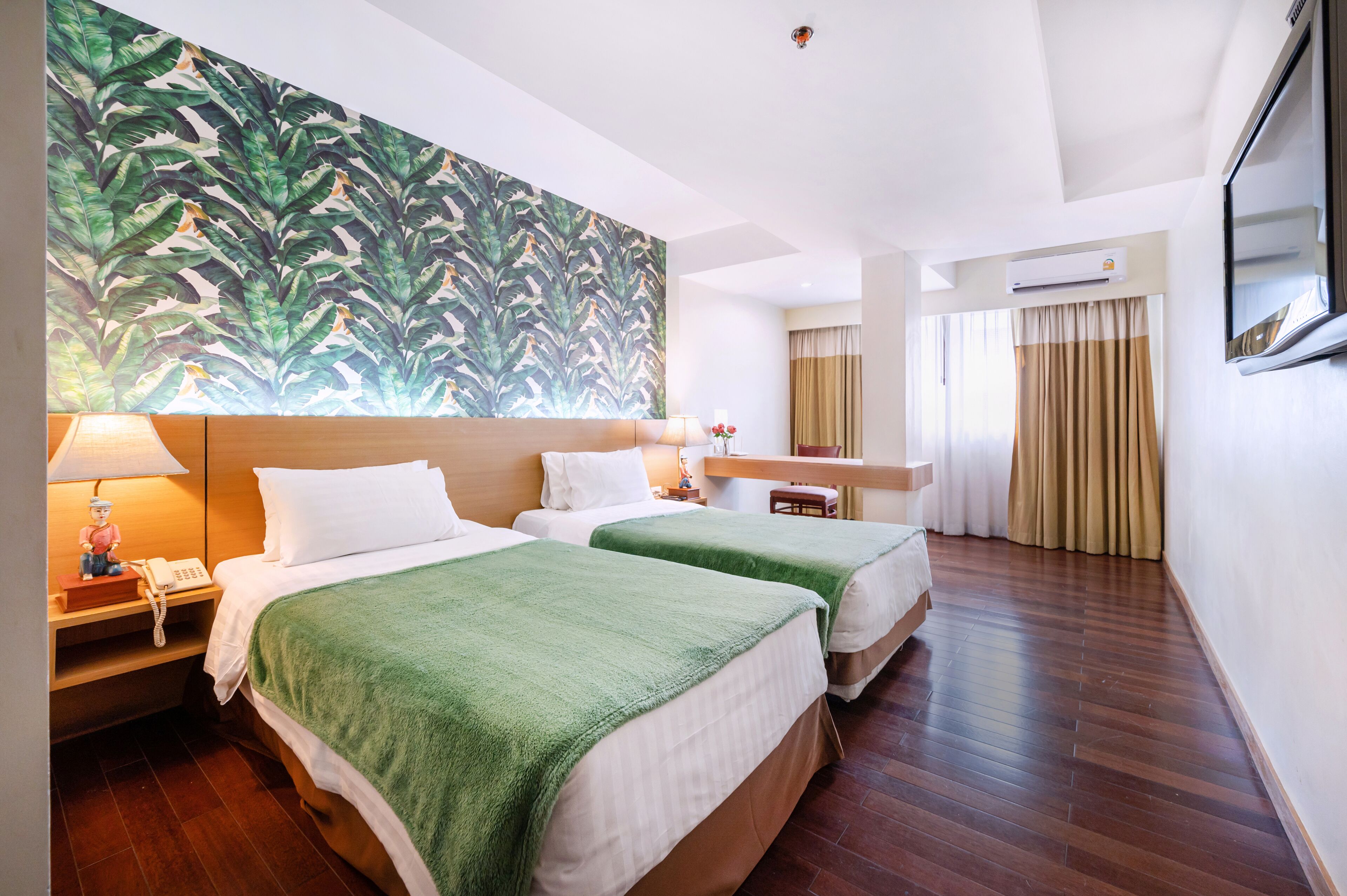 Habitación de hotel con dos camas, un cabecero de madera, una mesita de noche con lámpara, teléfono y una ventana con cortinas, además de una pared decorada con un estampado de hojas tropicales.