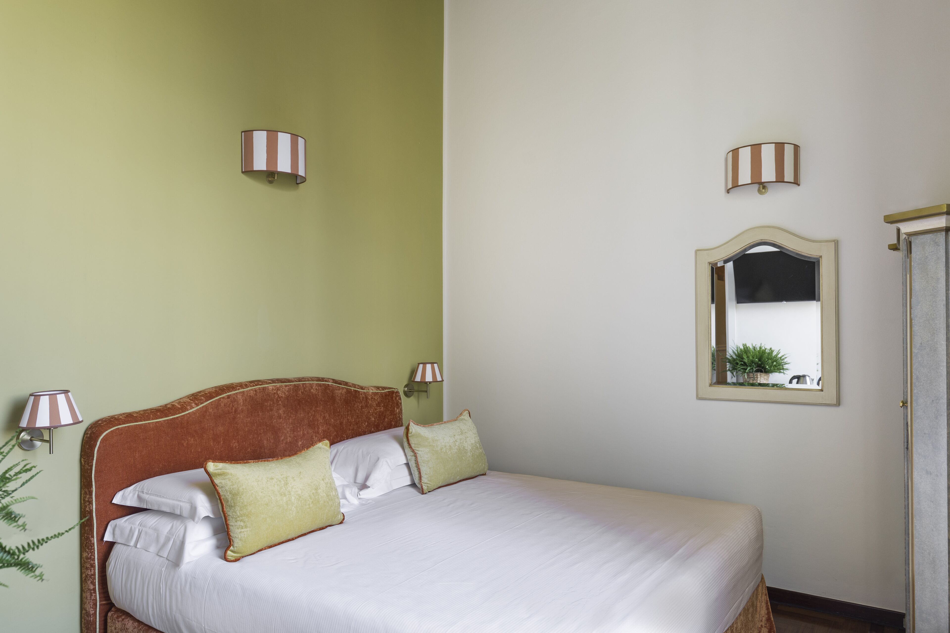 A neatly made bed with white linens and two pillows, a wall-mounted mirror, and two wall sconces with striped lamps.