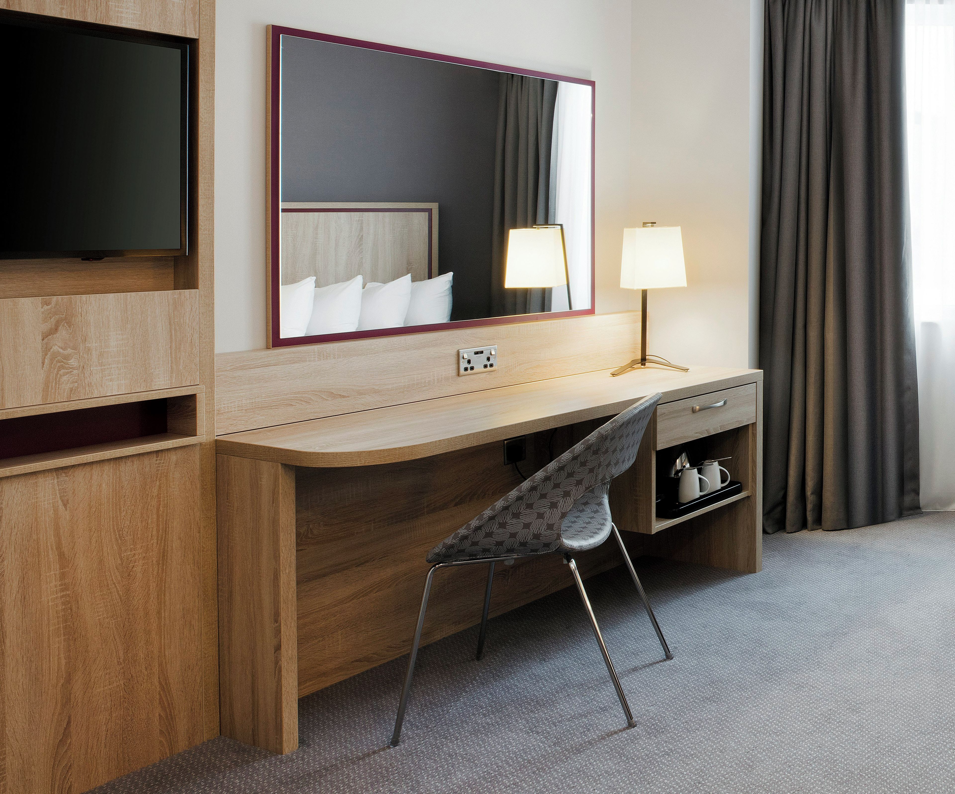 Een hotelkamer met een houten bureau, een stoel, een televisie, een spiegel, een lamp en een nachtkastje met een lamp.