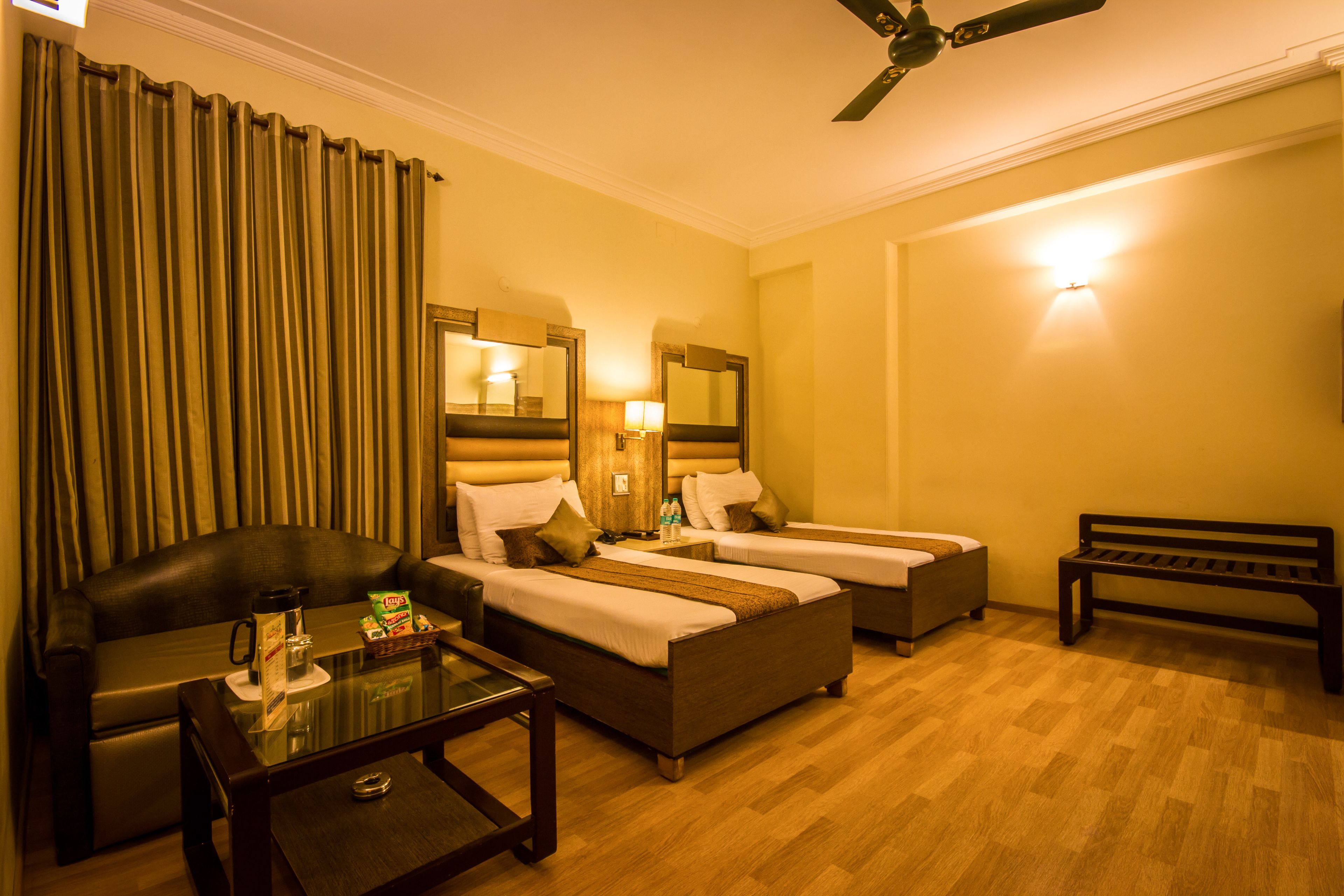 A hotel room with two single beds, a sofa, a coffee table, a bench, and a ceiling fan.
