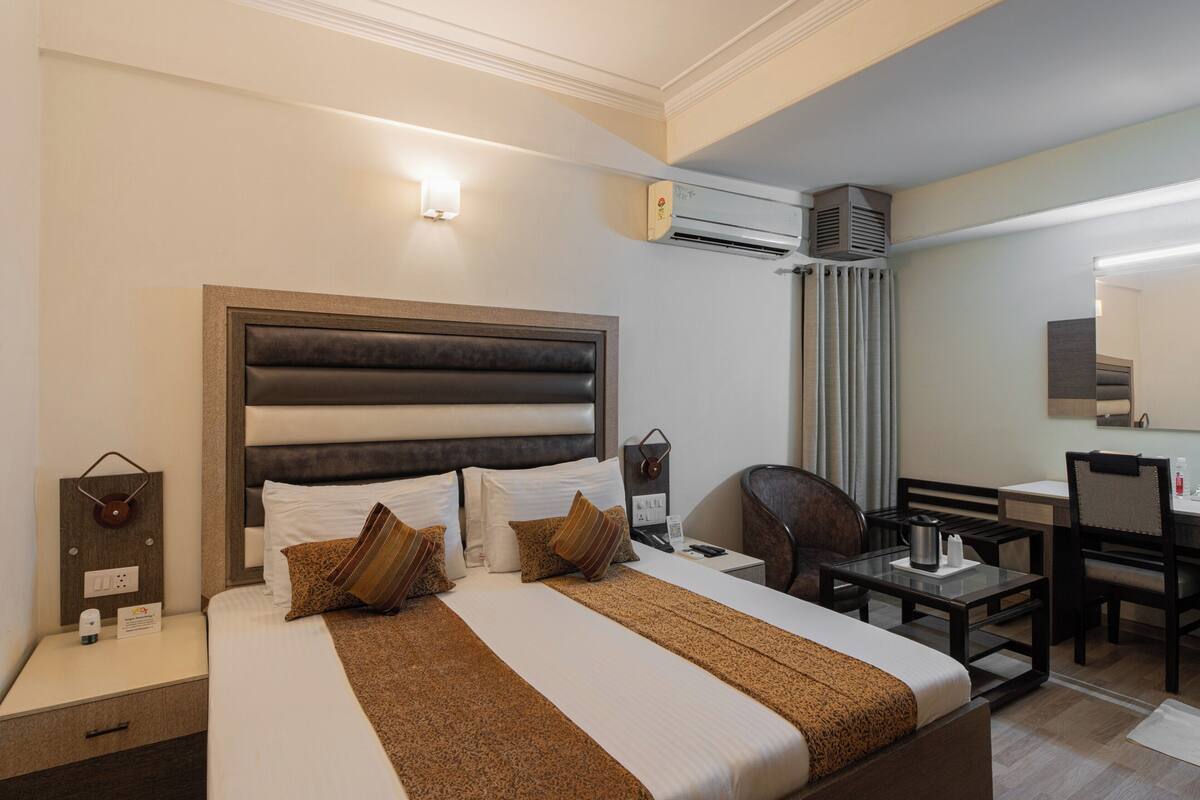 A hotel room with a large bed, bedside tables, a desk, a chair, an air conditioning unit, and a mirror.