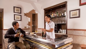 A bar area with a bartender, a customer, and a wine glass. There are framed pictures on the wall and a brick wall behind the bar. - Hotel Luciani (Rome)