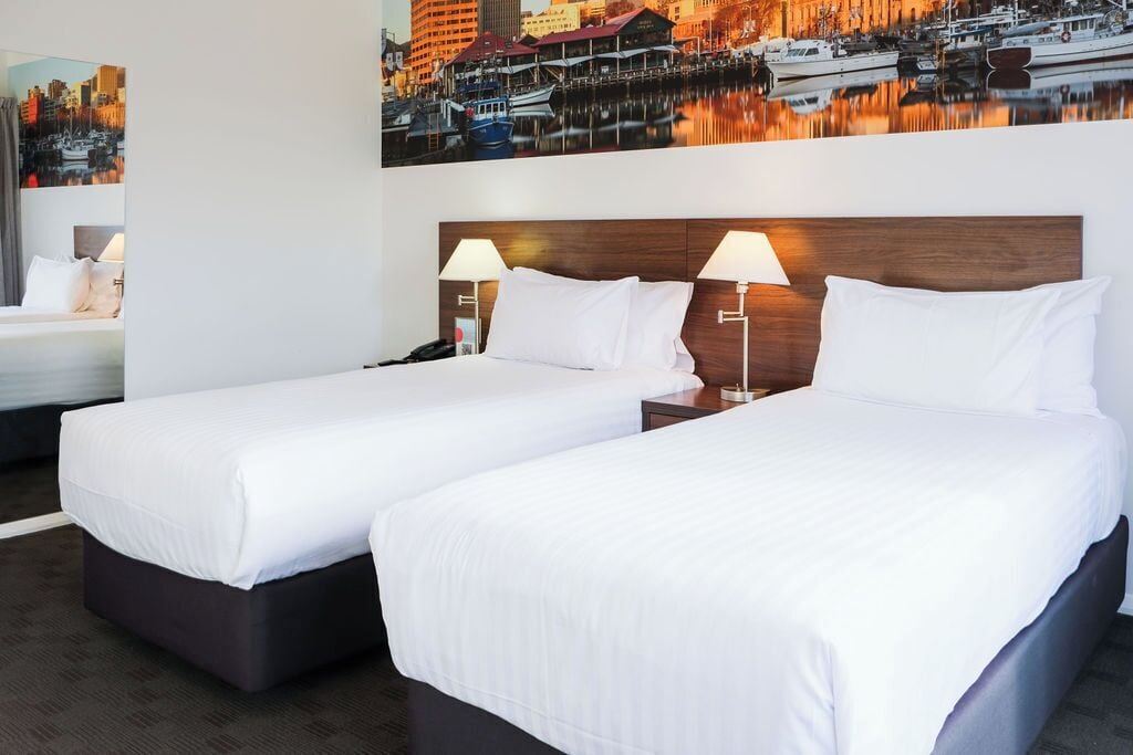 A hotel room with two beds, a nightstand with a lamp, and a large cityscape painting above the beds.