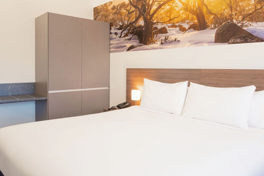 A hotel room with a bed, a wardrobe, a nightstand, and a wall-mounted lamp.