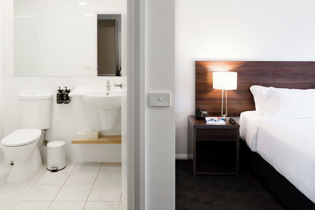 Une chambre d’hôtel comprenant un lit, une table de chevet avec une lampe, une petite commode, un miroir, des toilettes, un lavabo et une poubelle.
