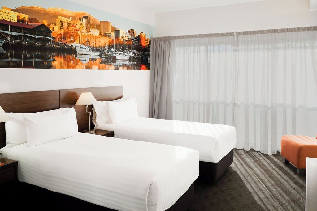A hotel room with two beds, a nightstand, a lamp, a window with curtains, and a cityscape mural on the wall.