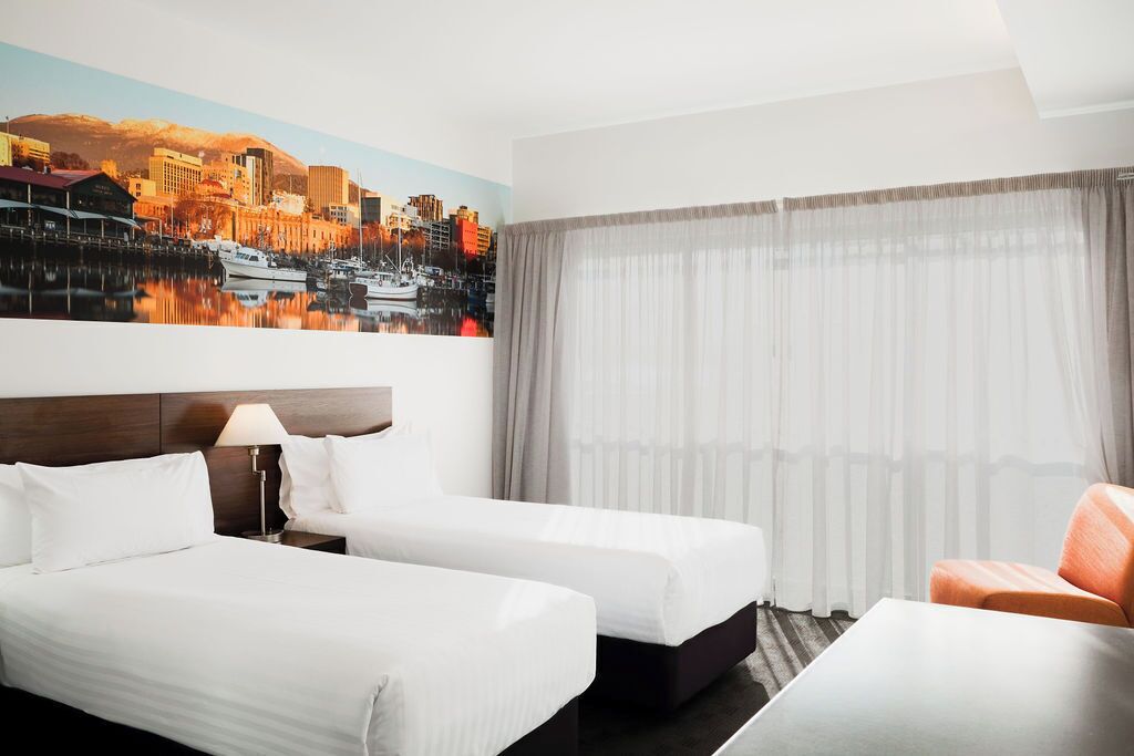 Une chambre d’hôtel avec deux lits, un canapé, un tableau représentant un paysage urbain et une grande fenêtre avec des rideaux.