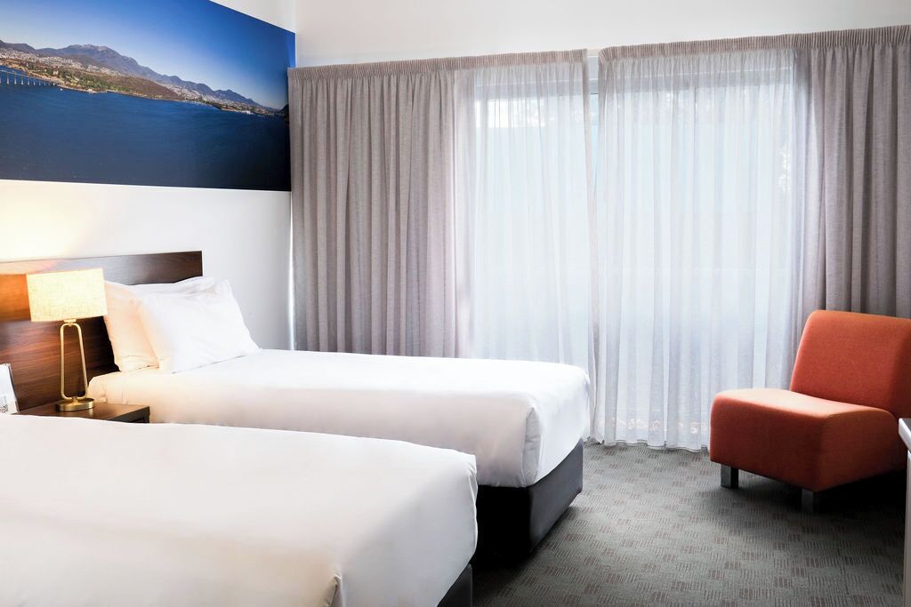 A hotel room with two beds, a chair, a lamp, and a view of a body of water through sheer curtains.