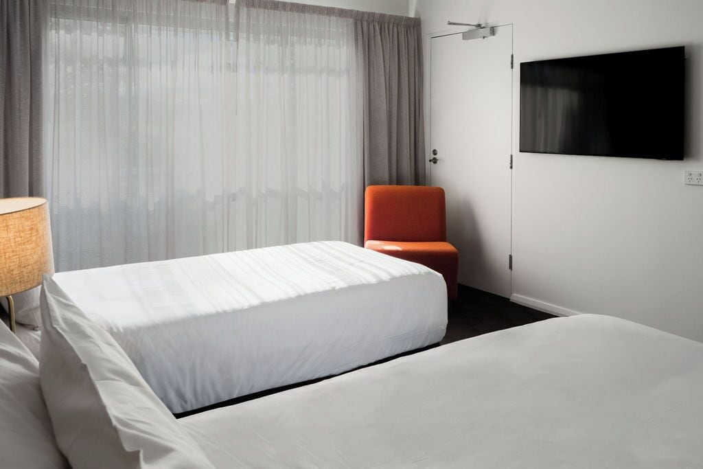 A hotel room with a bed, a chair, a TV on the wall, and a window with curtains.