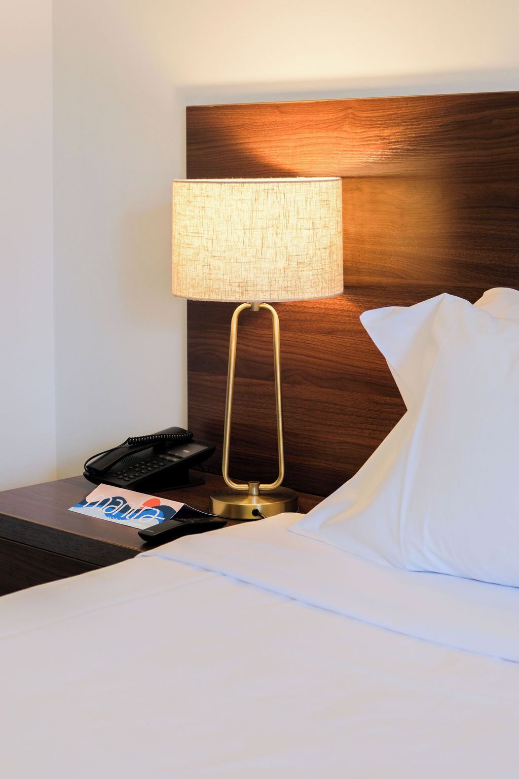 A hotel room with a bed, a nightstand with a lamp, a telephone, and a card.