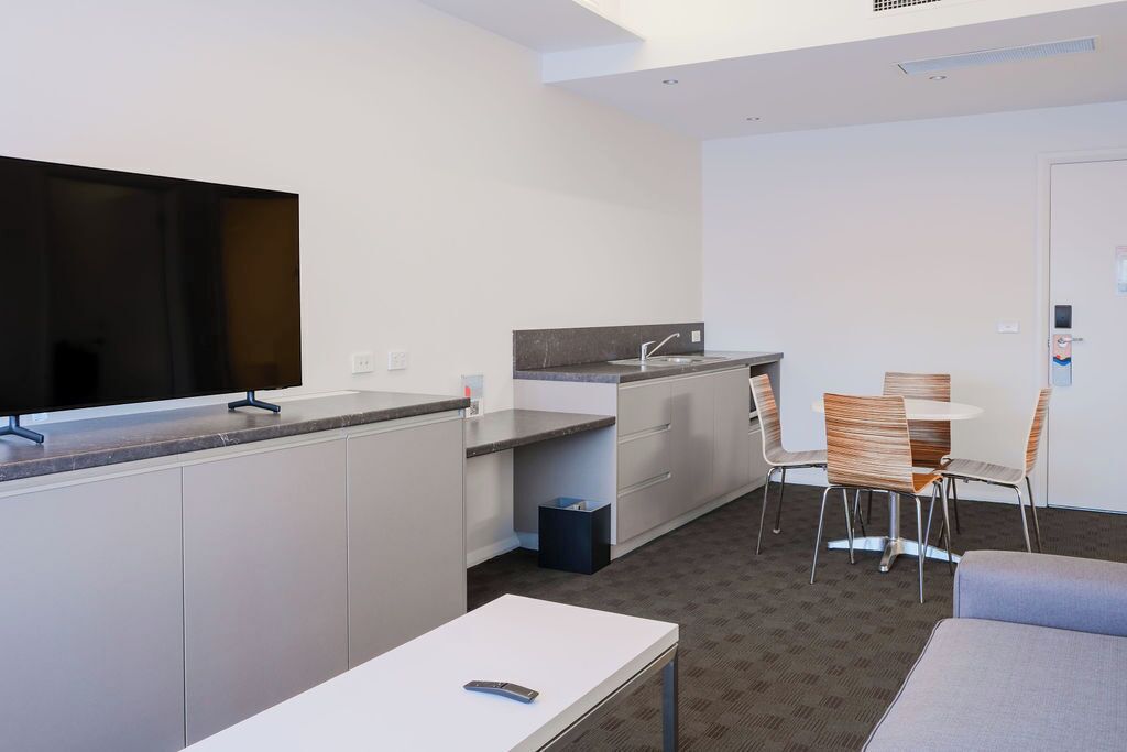 A compact hotel room with a kitchenette, a flat-screen TV, a dining area with chairs, and a sofa.