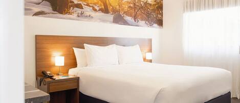 A hotel room with a large bed, a nightstand with a lamp, and a painting of a snowy landscape on the wall.