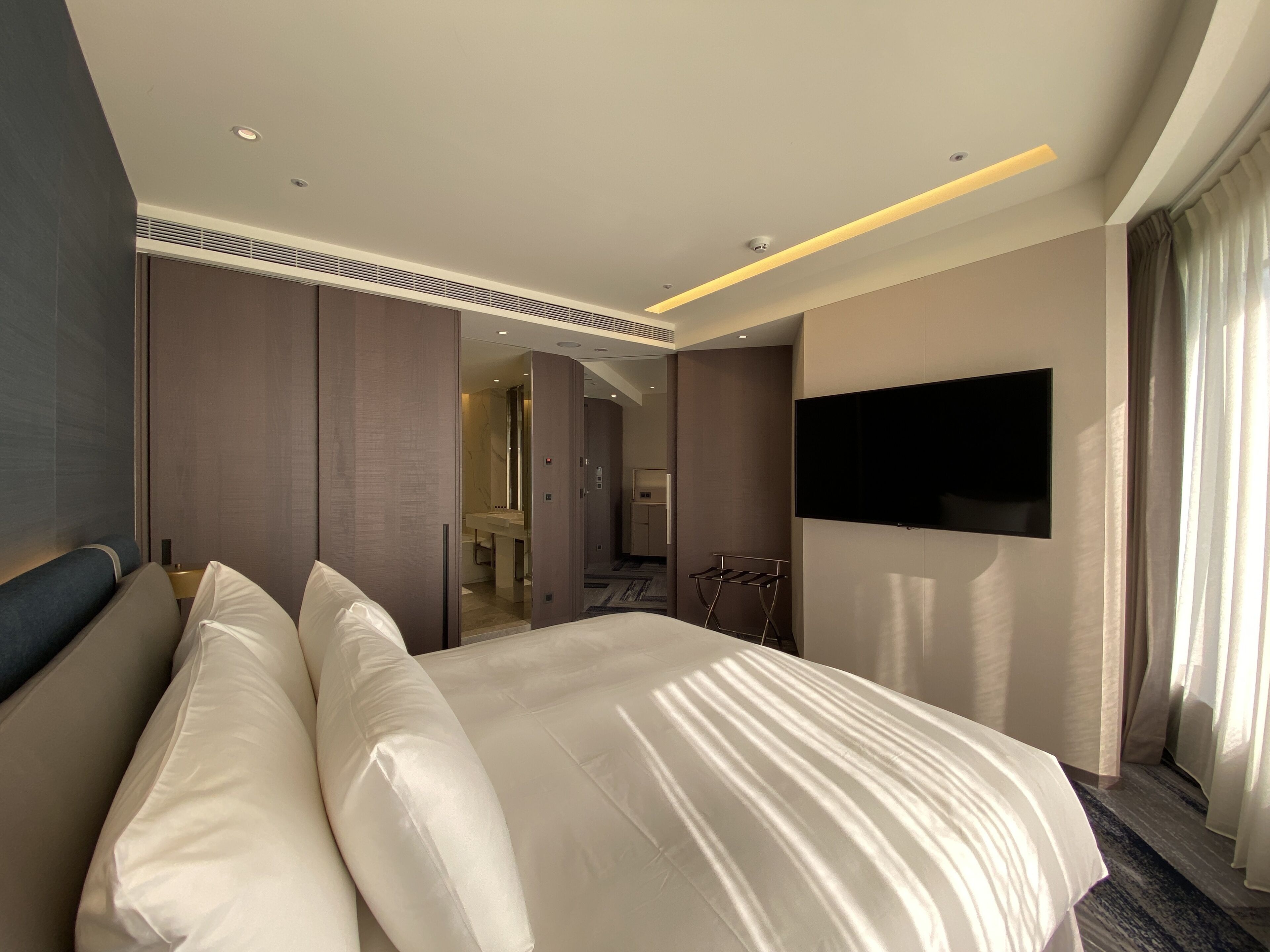 A modern hotel room with a large bed, a flat-screen TV mounted on the wall, and a bathroom visible through an open door.