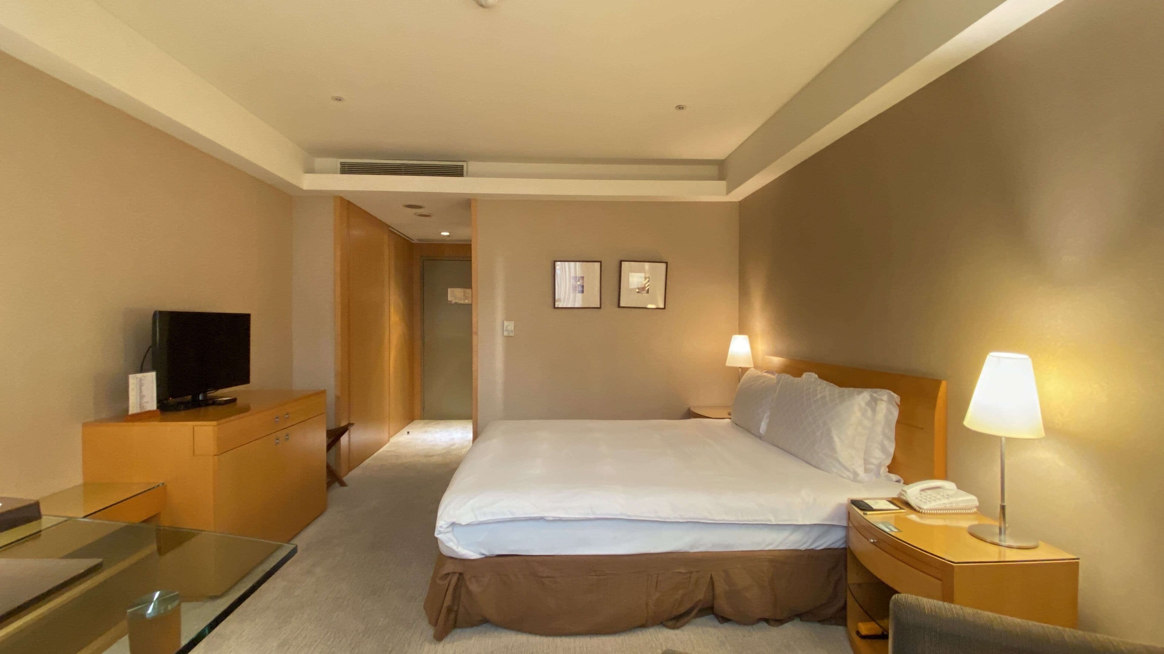 Una habitación de hotel con cama, lámparas de noche, un televisor sobre un aparador de madera y un escritorio con una silla.
