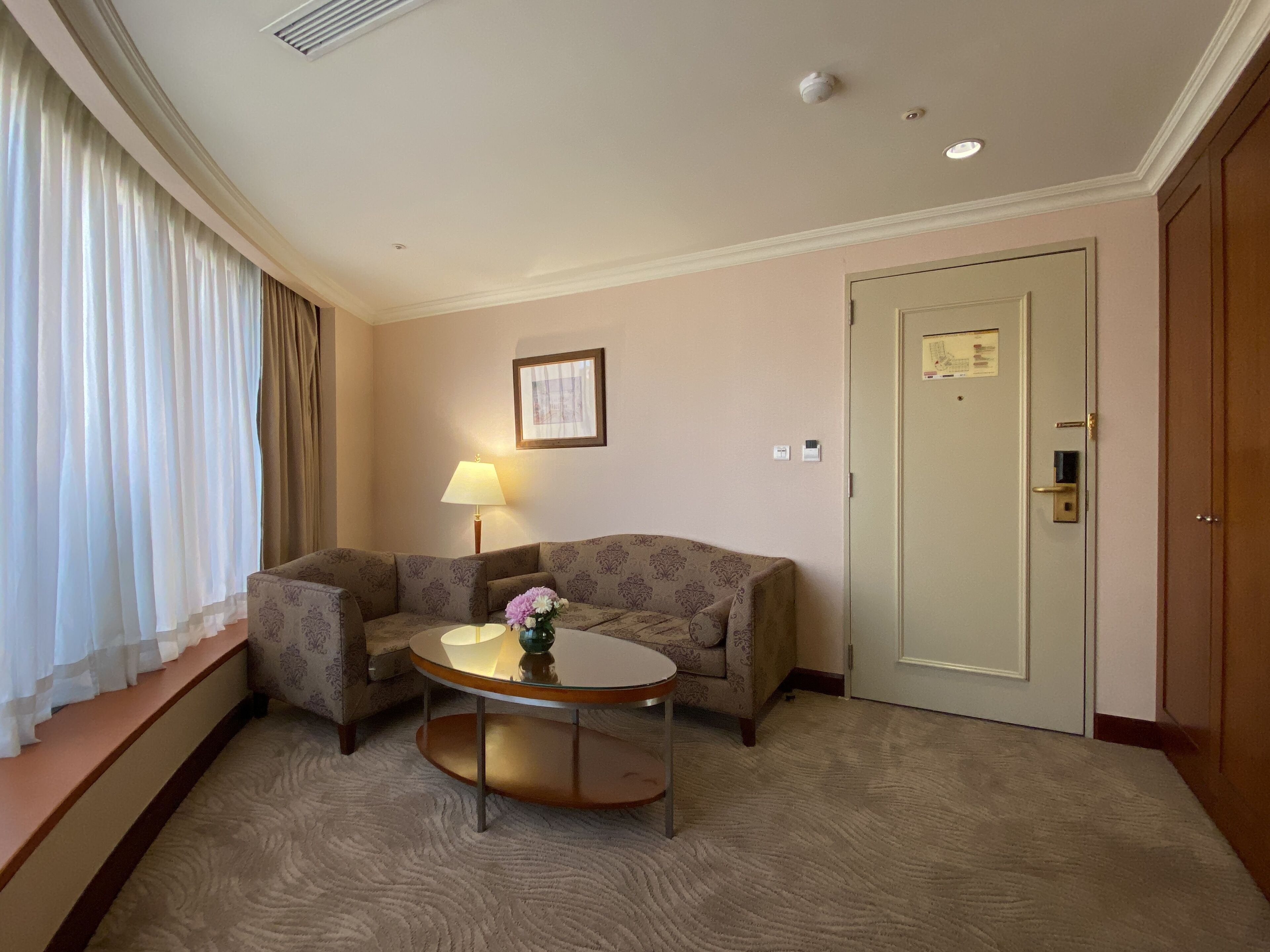 Una habitación de hotel con sofá, una mesita redonda con un jarrón, una lámpara y una puerta.