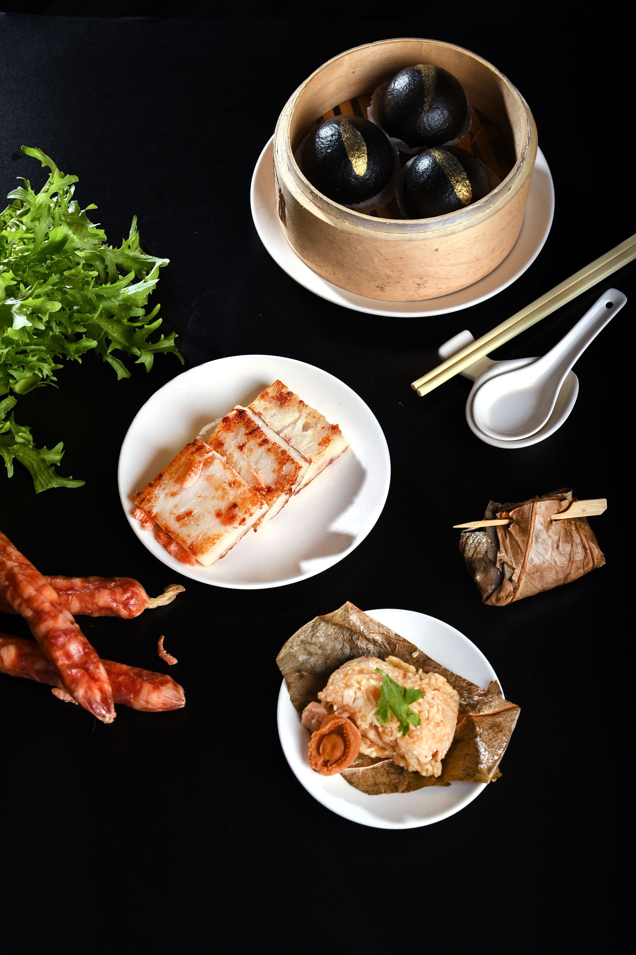 A table with various dishes, including a bowl of black and gold items, a plate with a slice of food, a wrapped item, and some vegetables.