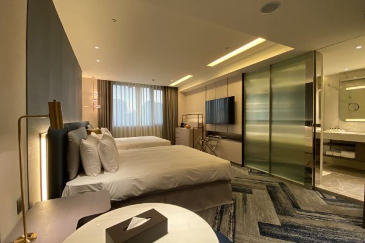Habitación de hotel moderna con cama, escritorio, televisión, baño con mampara de vidrio y vistas a la ciudad.