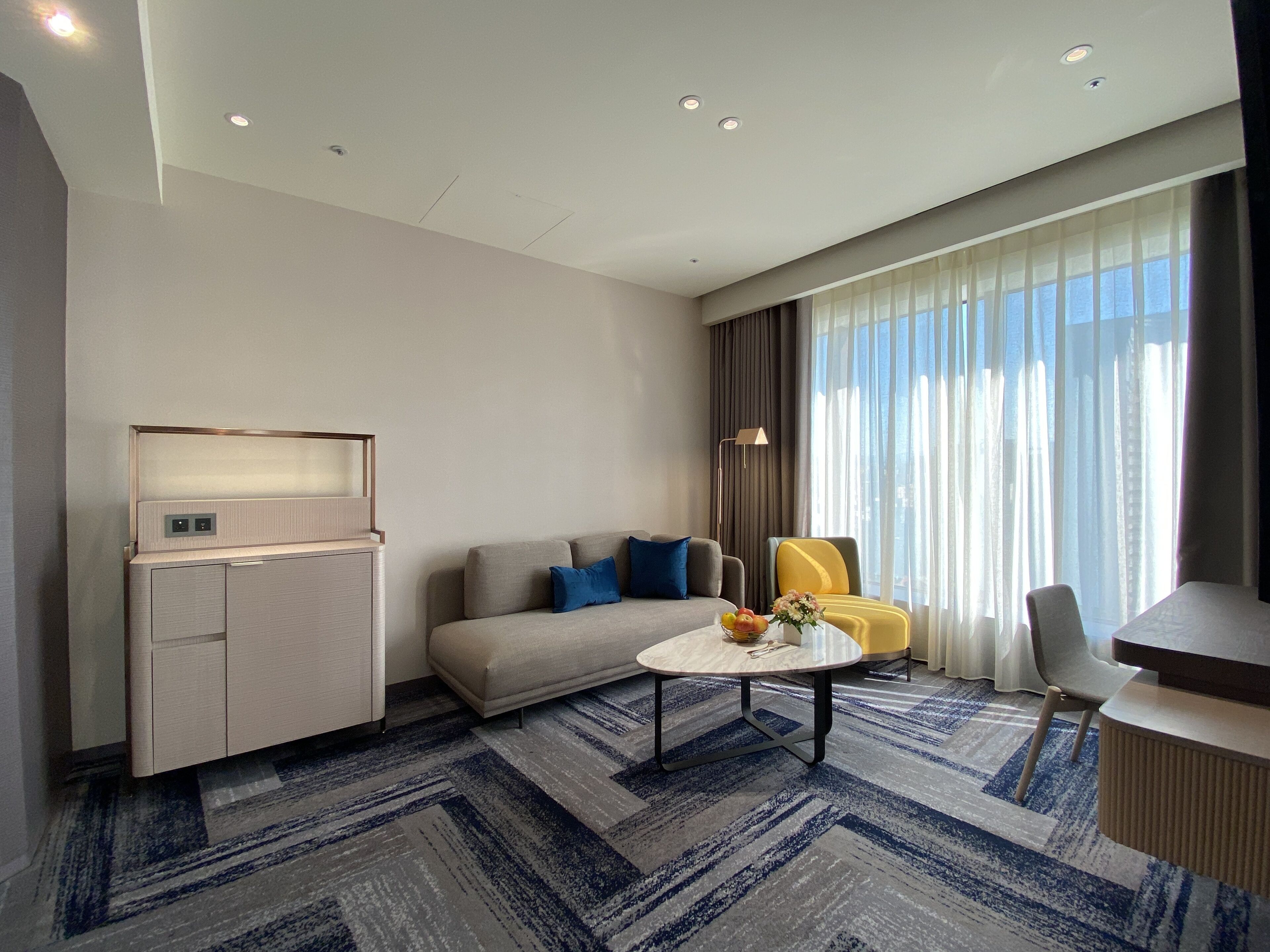 A modern hotel room with a sofa, a small table with a vase of flowers, a desk, and a large window with curtains.