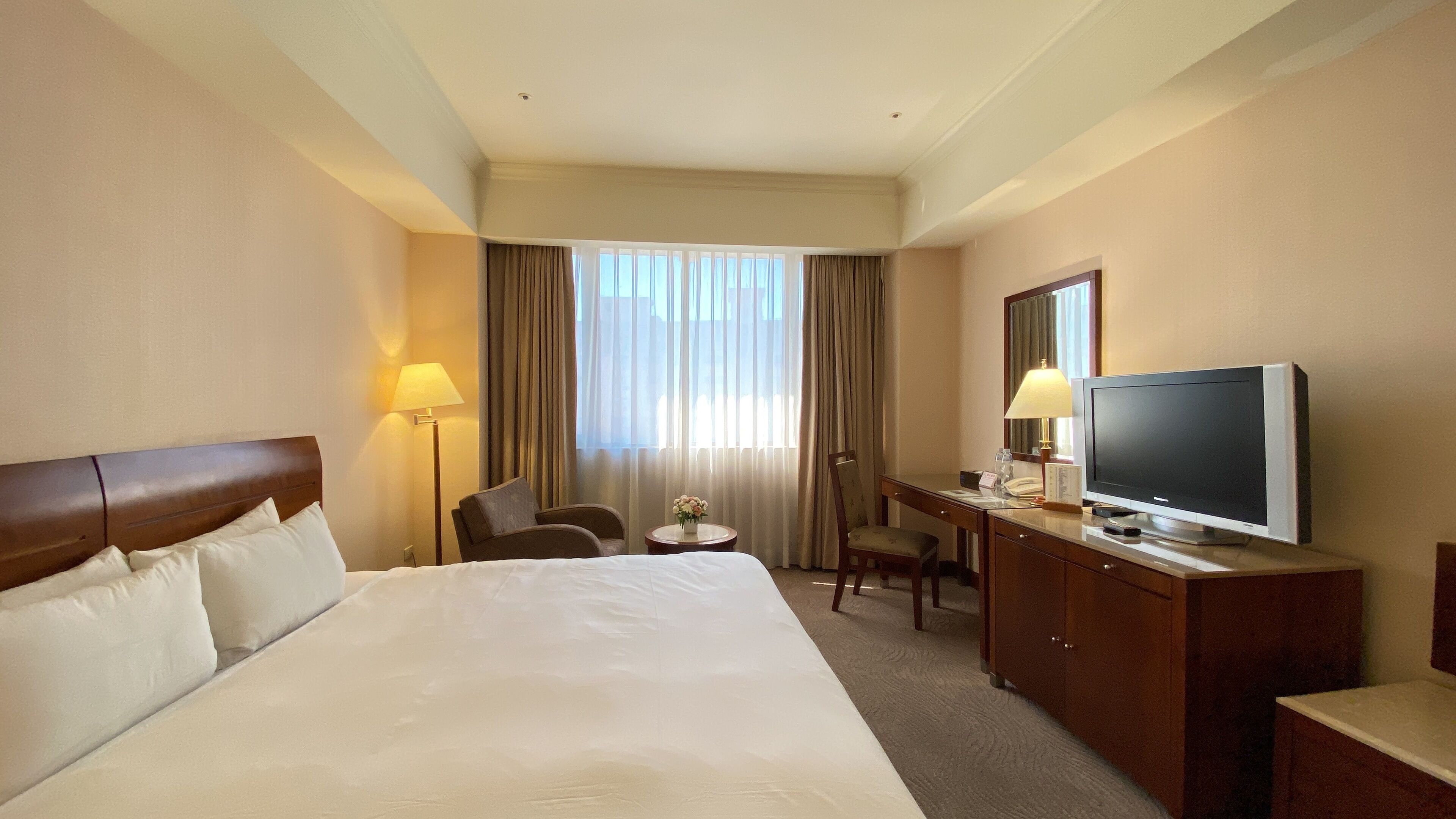 A hotel room with a large bed, a desk with a television, a chair, and a lamp.