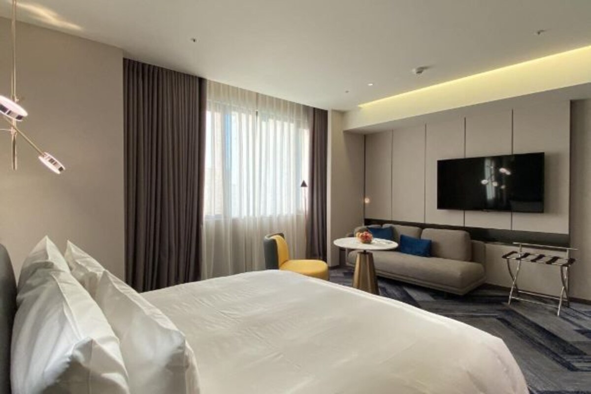 Una habitación de hotel con cama, sofá, una mesita redonda, televisor montado en la pared y ventana con cortinas.