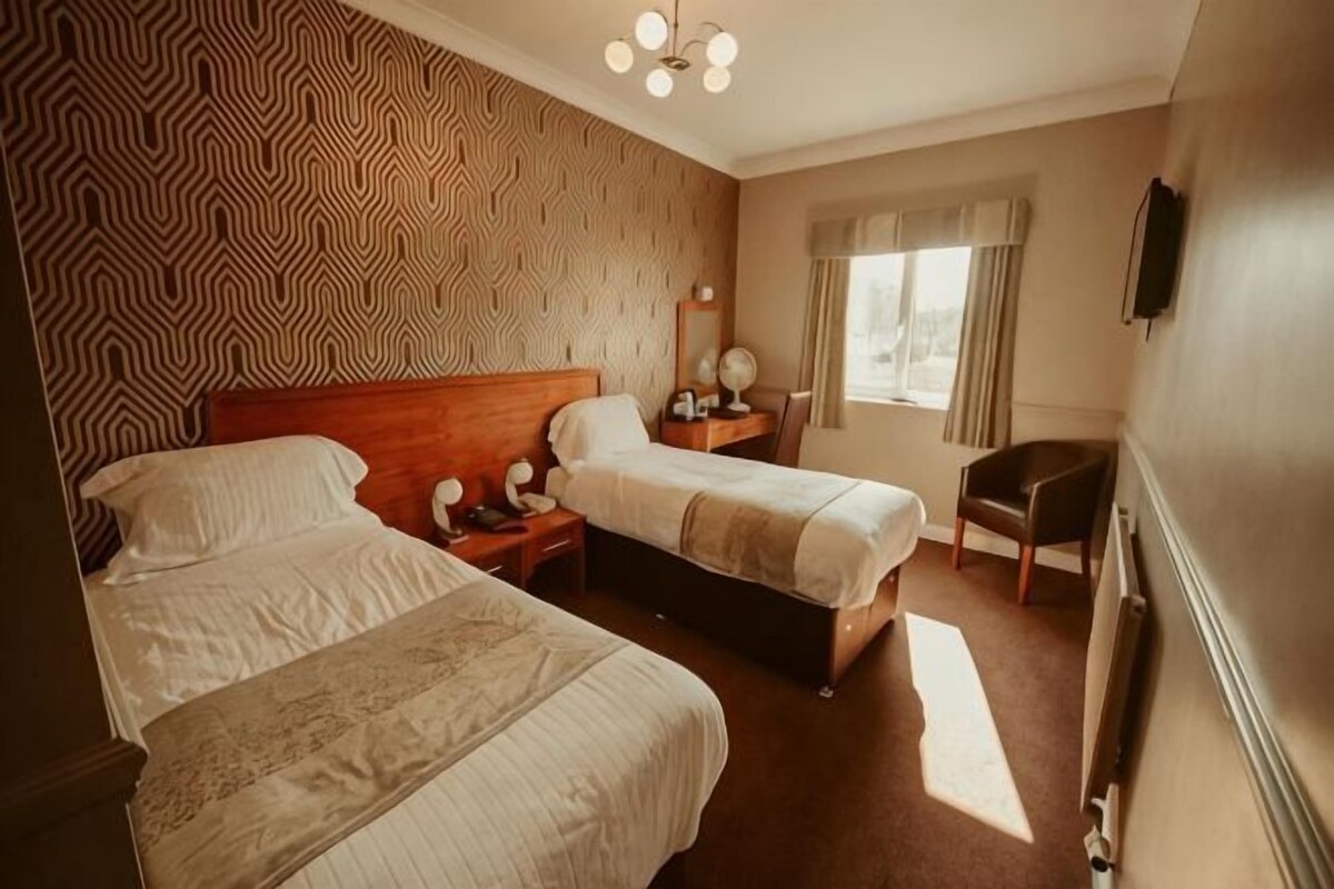 Habitación de hotel con dos camas, una mesita de noche, una silla y un ventanal con cortinas.