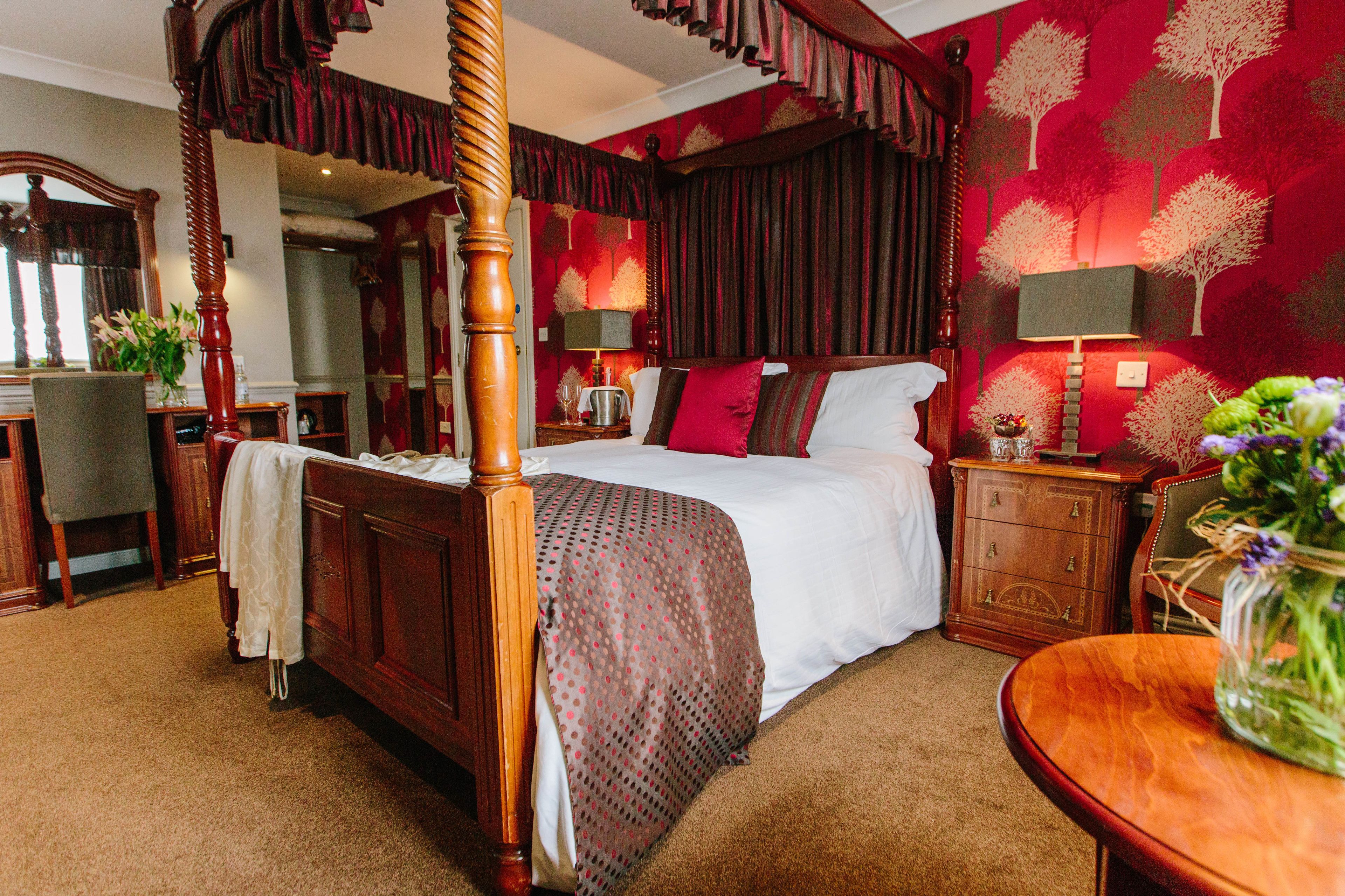 A four-poster bed with a white bedspread and red accents, a wooden nightstand with a lamp, and a vase with flowers on a wooden table.