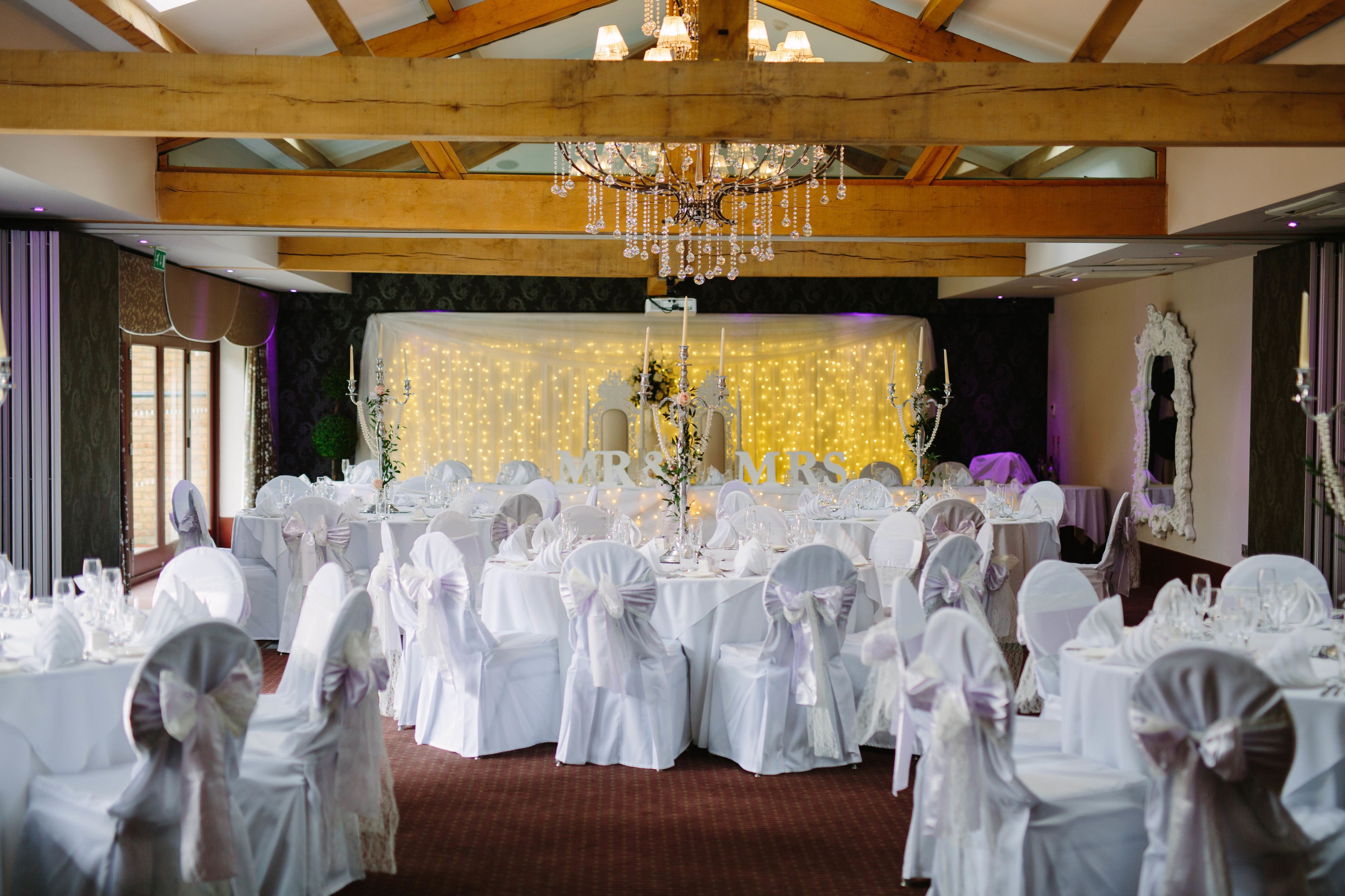 A banquet hall with round tables set for a wedding reception, featuring white tablecloths and chairs with bows.