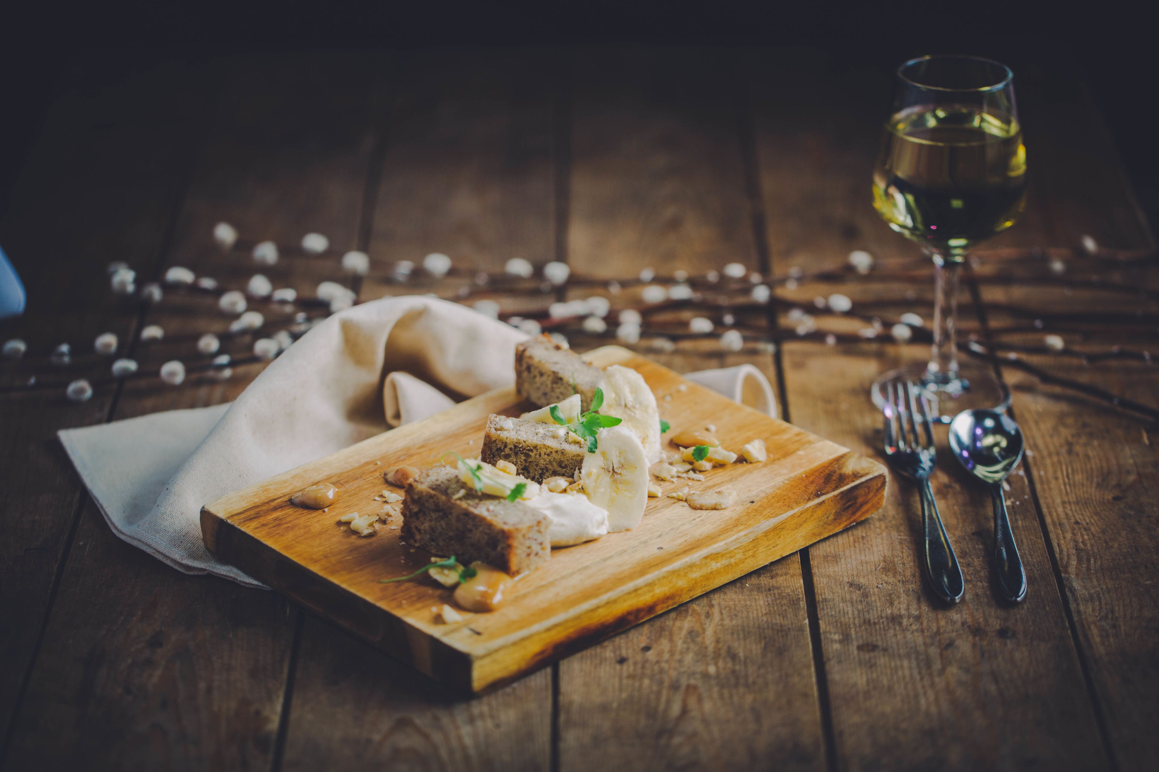 A wooden cutting board with slices of bread and cheese, a glass of white wine, and cutlery on a wooden table.