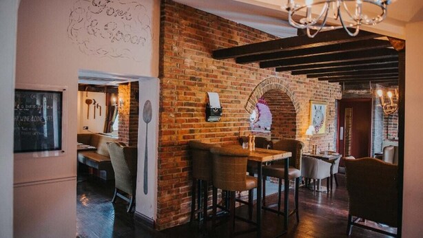 Un restaurante con paredes de ladrillo, mesas y sillas de madera. Hay un menú en una pizarra en la pared y una araña colgando del techo.